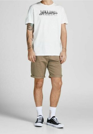 Hvid grafisk t-shirt med sort tekst, beige rullede shorts, hvide sokker med mønster og sorte sneakers. Neutral baggrund.