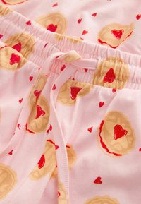 Un pantalon de pyjama rose à taille élastique avec cordon présente un motif de tartes en forme de cœur et de petits cœurs rouges sur un tissu doux.