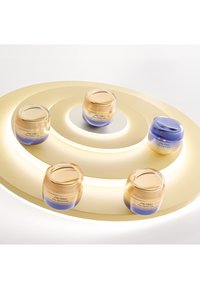 Fem krämburkar med guld- och lila lock, märkta "Shiseido Vital Perfection", arrangerade på en guldrund bas med belysta accenter.