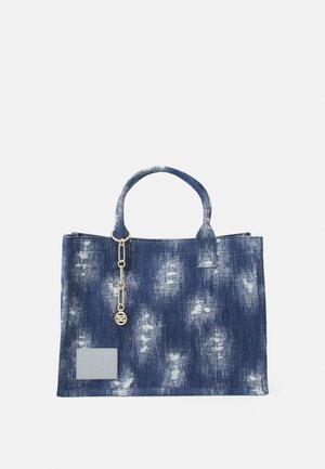 Denim shopper med håndtag, slidte hvide lapper, en guldkæde charms og en firkantet syet lap nederst til venstre.