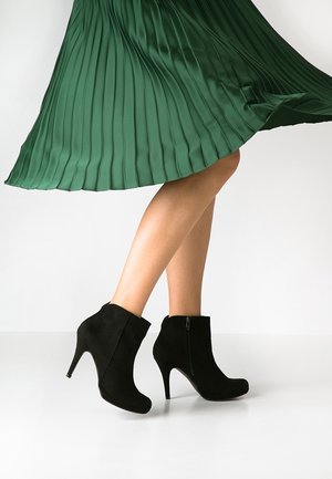 Femme portant des bottines noires à talons hauts et une jupe plissée verte fluide, debout sur un sol blanc, jambes croisées.