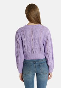 Lila cable-knit pulóver, bordázott kerek nyakkal és bonyolult mintázattal a háta részén. Kék farmernadrággal viselve.