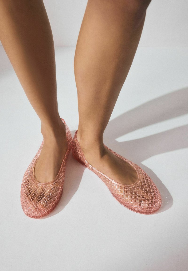Transparente rosa Flats mit einer gemusterten Oberfläche und offenen Seiten, die ein geometrisches Design und eine glatte Textur zeigen.