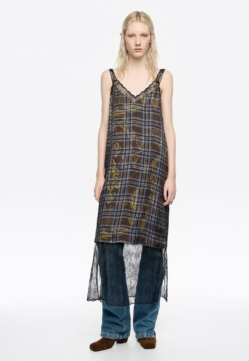 Vestido largo de cuadros con ribete de encaje. Fondo oscuro con líneas amarillas y azules, superpuesto por encaje y pantalones de mezclilla azul acampanados.