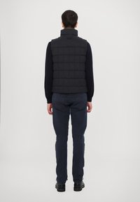Black quilted vest with a high collar, shown from the back. Paired with navy pants and black shoes. Simple design, no visible hardware.