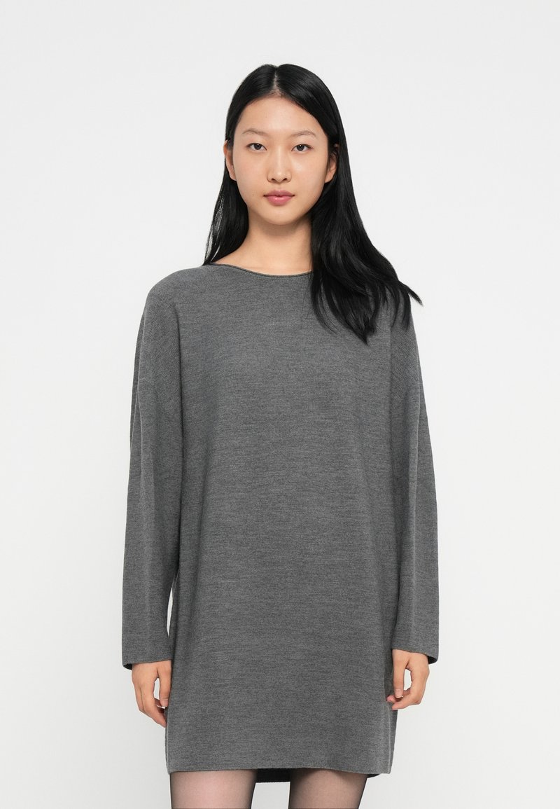 Giovane donna con lunghi capelli neri, che indossa un vestito in maglia grigio a maniche lunghe e larghe, in piedi contro uno sfondo bianco semplice.