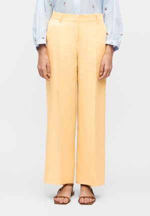 Personne portant un pantalon taille haute, jambes larges, couleur pêche clair, une chemise rayée bleue avec broderie florale, et des sandales ouvertes marron, debout.