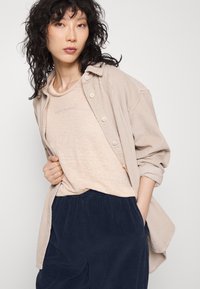 Heled beige soonist nööbideta särk heleda beeziga särgi peal, koos tumesiniste sametpükstega. Omab pehmeid tekstiile ja mugavat lõiget.