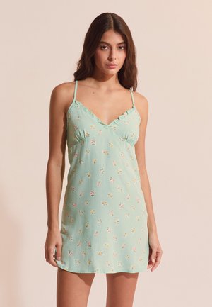 Mujer joven con un vestido de tirantes fino de flores en verde menta, posando frente a un fondo beige liso.