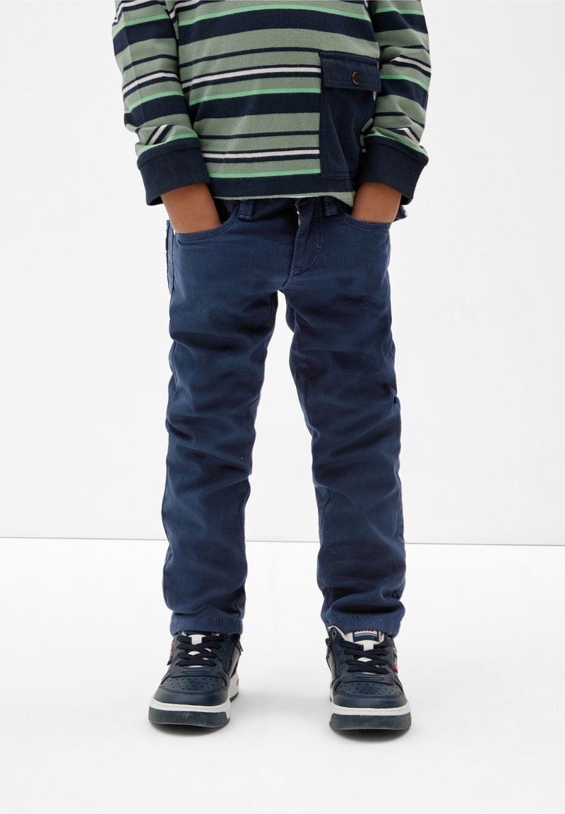 Enfant portant un pantalon bleu marine, des baskets bleu marine et un shirt à manches longues rayé vert et bleu marine, les mains dans les poches, debout sur un sol blanc.