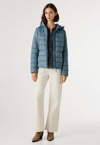 Jonge vrouw in een lichtblauwe pufferjas, met een donkere gedessineerde blouse, off-white wijde broek en bruine enkellaarzen, staand tegen een witte achtergrond.