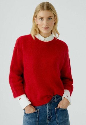 Roter Wollpullover mit Rundhalsausschnitt und lockerer Passform. Darunter wird ein weißes Hemd mit Kragen getragen, das rote Lippenmuster an den Ärmeln aufweist. Dazu werden Jeans aus Denim getragen.