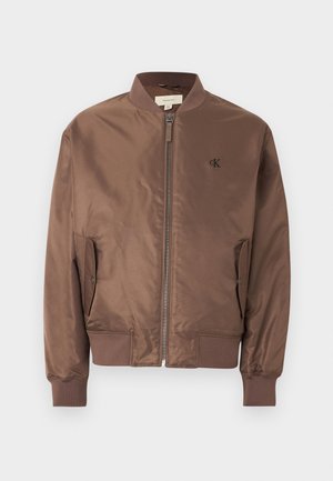 Ruda bombonešio striukė su priekiu užtrauktuku, gofruotu apykakle, rankogaliais ir apačia, šoninėmis kišenėmis su atvartais ir mažyčiu „cK“ logotipu ant kairio krūtinės.