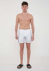 Pantaloni da bagno bianchi con vita regolabile, caratterizzati da strisce laterali nere con testo. Indossati con sandali neri a ciabatta, il modello è in piedi dritto.