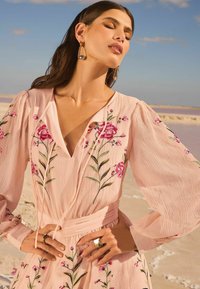 Lichtroze jurk met bloemmotieven, lange mouwen en een ceintuur om de taille. Model draagt gouden oorbellen en een ring, tegen een achtergrond van het strand.