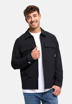 Uśmiechnięty młody mężczyzna z kręconymi włosami, ubrany w czarną kurtkę na zamek błyskawiczny, pod którą ma białą koszulę, oraz niebieskie dżinsy, na jasnym tle.