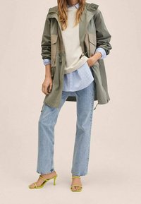 Parka vert et beige avec une capuche, superposée à un pull blanc et une chemise à rayures bleues, associée à un jean bleu clair et des sandales à talons vertes.