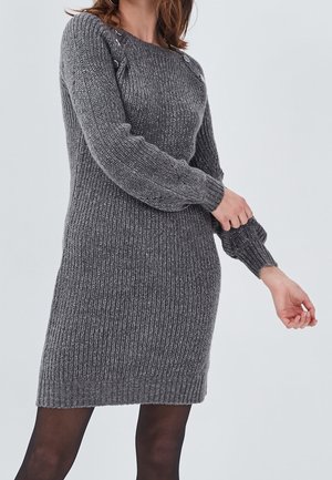 Robe-pull grise à manches longues en tricot avec des boutons sur l'épaule, portée par une personne ajustant la manche, sur un fond clair uni.