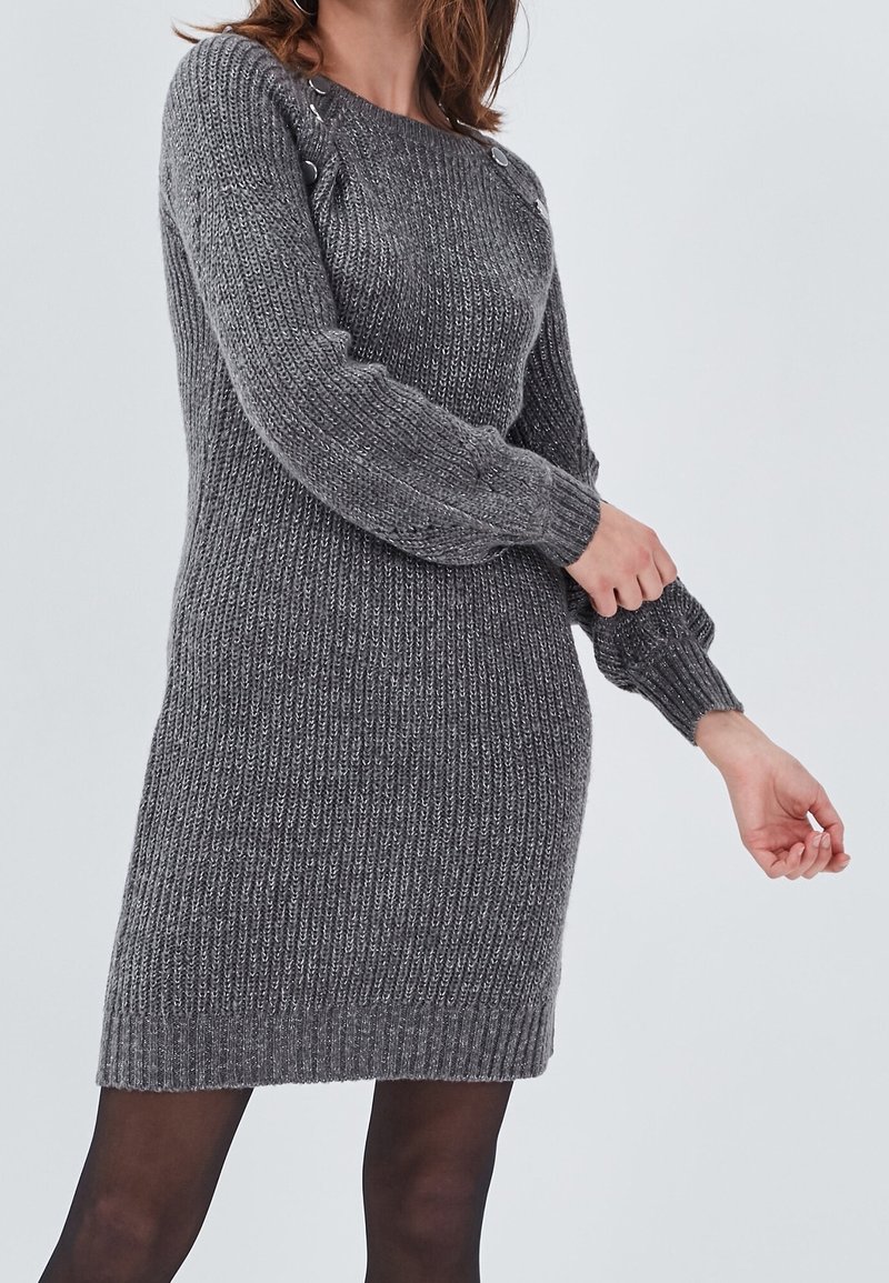Robe-pull grise à manches longues en tricot avec des boutons sur l'épaule, portée par une personne ajustant la manche, sur un fond clair uni.