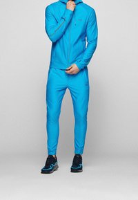 Survêtement pour homme en tissu bleu vif ; veste à capuche zippée avec logo, pantalon slim avec poches latérales, chaussures accentuées en bleu foncé.
