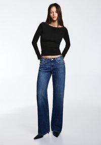 Zwarte top met lange mouwen gecombineerd met een high-waisted, wijdvallende blauwe spijkerbroek. De spijkerbroek heeft subtiele stiksels en voorzakken. Zwarte puntige schoenen.