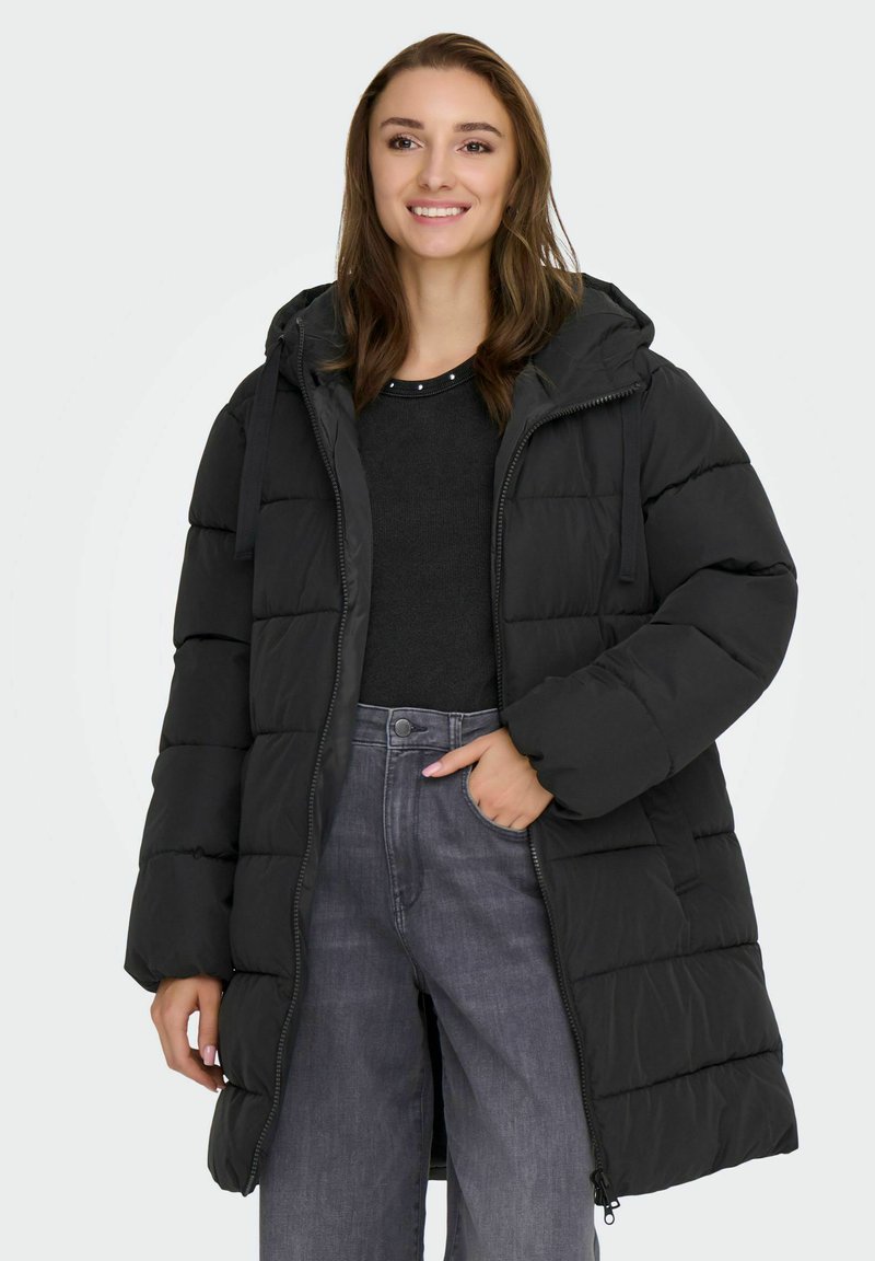 Melna pufota jaka ar kapuci, ar quiltētām daļām un rāvējslēdzēja aizdari, valkāta virs tumša krekla un džinsiem. Mīksta tekstūra un plaša piegriezuma.