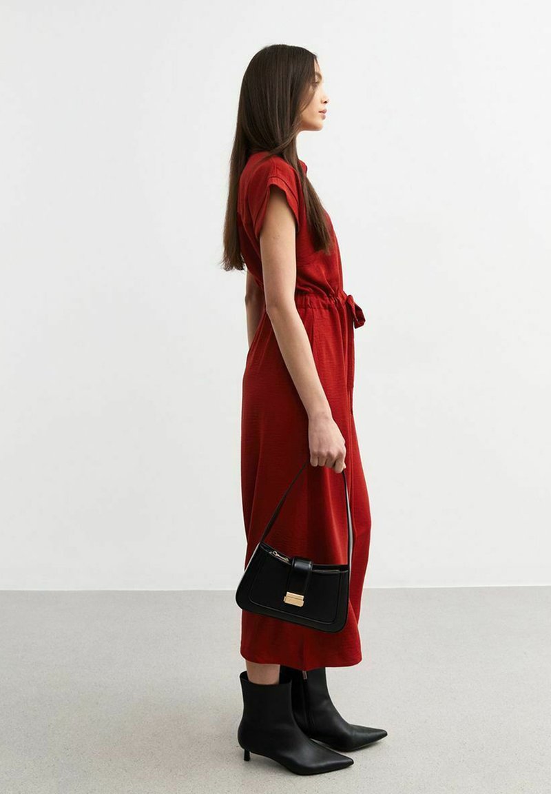 Vestido rojo, hasta la rodilla, con mangas cortas y cinturón anudado. Combinado con botas negras hasta el tobillo y un pequeño bolso negro con herrajes dorados.