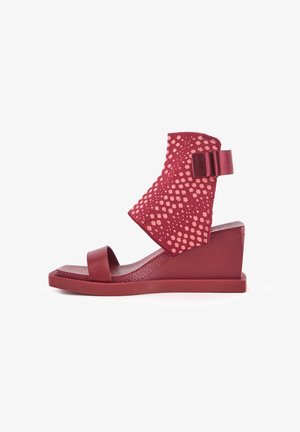 Sandale compensée rouge avec une tige texturée à motif floral, une lanière lisse sur les orteils et un large accent décoratif à l'arrière.