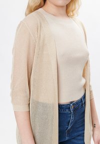 Cárdigan de punto beige claro con mangas tres cuartos, de frente abierto y una textura suelta y aireada, sobre una blusa de punto crema y jeans de mezclilla azules.