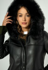 Zwarte leren jas met een grote nepbont capuchon. Heeft een ritsluiting en een getailleerd ontwerp. Binnenvoering zichtbaar met een groen sweaterdetail.