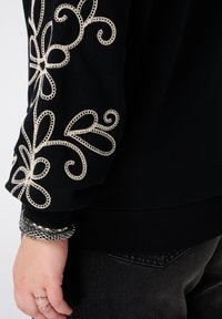 Zwarte trui met ingewikkelde beige bloemenborduursels op de mouwen. Voorzien van een geribbelde zoom en manchetten. Zilveren metallic armbanden zichtbaar om de pols.