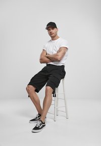 Melni kargo šorti, balta T-kreklu un melna cepure, sēž uz baltā krēsla. Melni sporta apavi ar baltām detaļām. Gludas tekstūras, ikdieniška dizains.