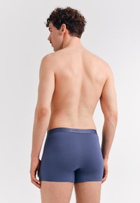 Young man with curly hair stands facing away, wearing fitted navy blue boxer briefs against a plain light background.