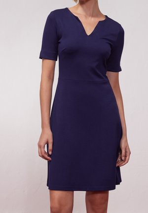 Vrouw draagt een knielange, kortermige donkerblauwe jurk met een V-hals met inkeping tegen een effen lichte achtergrond.