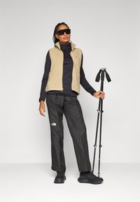 Beige fleece vest over een zwart langemouwen top, gecombineerd met zwarte waterdichte broek en zwarte wandelschoenen, met trekkingstokken in de hand.
