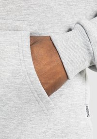 Grauer Pullover mit einer Fronttasche. Der Stoff ist weich und hat eine glatte Textur. Verfügt über gerippte Bündchen und ein Etikett an der Seite.