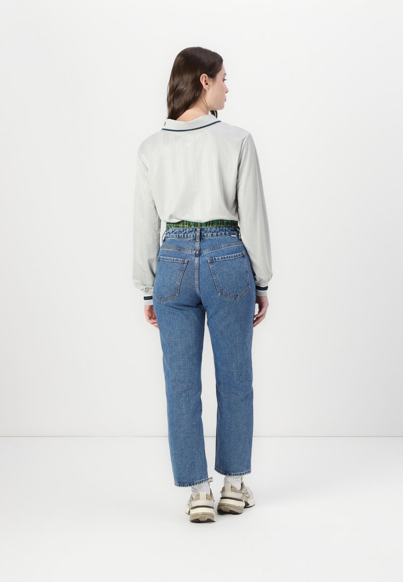 Camisa de manga larga gris claro con cuello, combinada con jeans de tiro alto azules y zapatillas blancas gruesas. Cinturilla con un patrón verde visible.