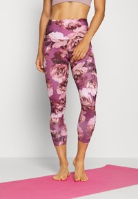 Osoba ubrana w fioletowe legginsy w kwiatowy wzór i jasnoróżowy top na krótko, stojąca boso na różowej macie do jogi.