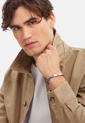 Giacca di cotone beige con chiusura a bottoni, indossata sopra una t-shirt bianca. Un bracciale metallico con scanalature orizzontali è visibile sul polso.