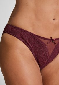 Bordeauxfarbene Spitzenunterwäsche mit floralen Mustern, transparenten Mesh-Elementen und einem kleinen Schleifendetail mit einem dekorativen Edelstein in der Mitte vorne.