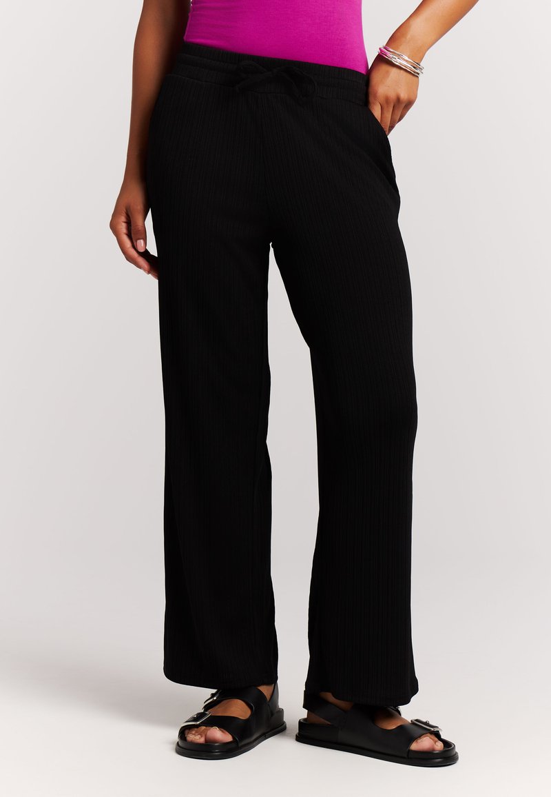 Schwarze, mit Nadelstreifen versehene Hose mit lockerem Schnitt, Kordelzugbund und Seitentaschen. Kombiniert mit flachen schwarzen Sandalen.