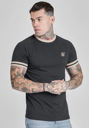 Homme avec des tatouages sur les bras et le cou, portant un t-shirt noir ajusté avec des bordures beiges et un jean bleu clair, regardant sur le côté avec les mains croisées.
