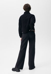 Zwarte denimjas met kraag, gecombineerd met hooggetailleerde zwarte wijde broekspijpen en grove zwarte schoenen. Gladde stof, minimale details.