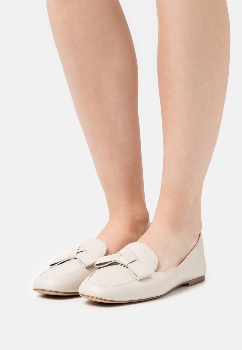 Mocassins en cuir beige clair avec un bout arrondi et un détail nœud à l'avant. Semelle plate, texture lisse et design minimaliste.