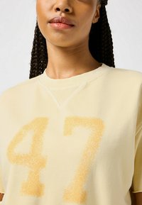 Felpa gialla chiara con una grande stampa testurizzata arancione "47". Il tessuto ha una consistenza morbida e scollo rotondo.