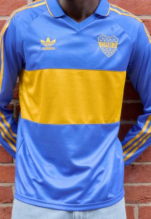 Personne portant un maillot de sport Adidas manches longues bleu et jaune avec logo "CABJ" debout contre un mur en briques rouges.