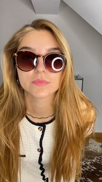 Óculos de sol redondos com armação em tartaruga, lentes escuras e acabamento brilhante. O modelo tem cabelo longo e ondulado e está a usar uma blusa branca texturizada.