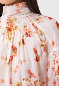 Witte bloes met bloemenprint, gekenmerkt door zachte plooien, een hoge hals en levendige oranjerode en roze bloempatronen op lichte stof.