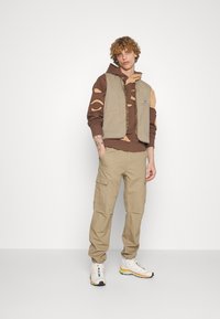 Beige ärmlös väst över en brun mönstrad hoodie, kombinerad med ljusbruna cargobyxor och vita sneakers med färgglada accenter.