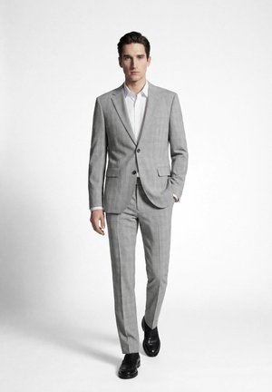 Mann in hellgrauem kariertem Anzug mit weißem Hemd und schwarzen Schuhen geht vor einem schlichten weißen Hintergrund nach vorne.